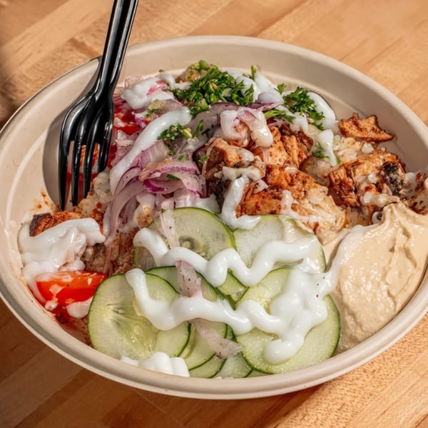 A Nana Street Food bowl with chicken, rice, hummus, cucumbers, tomatoes, red onions, and white sauce.