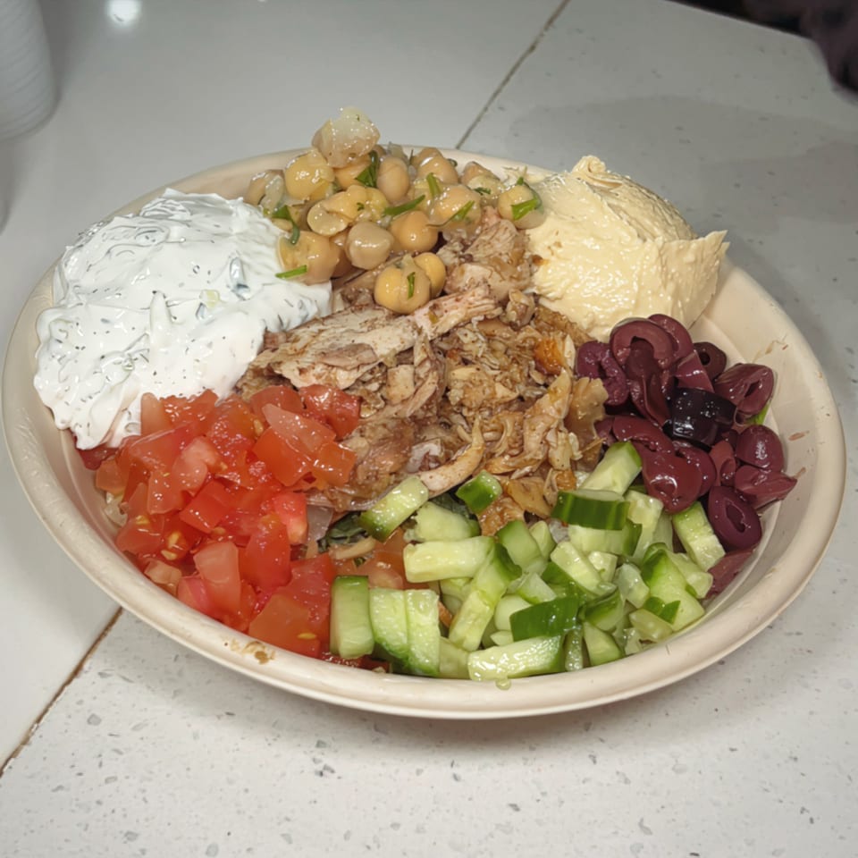 A Nana Street Food bowl with chicken, hummus, tzatziki, chickpeas, tomatoes, cucumbers, and olives.